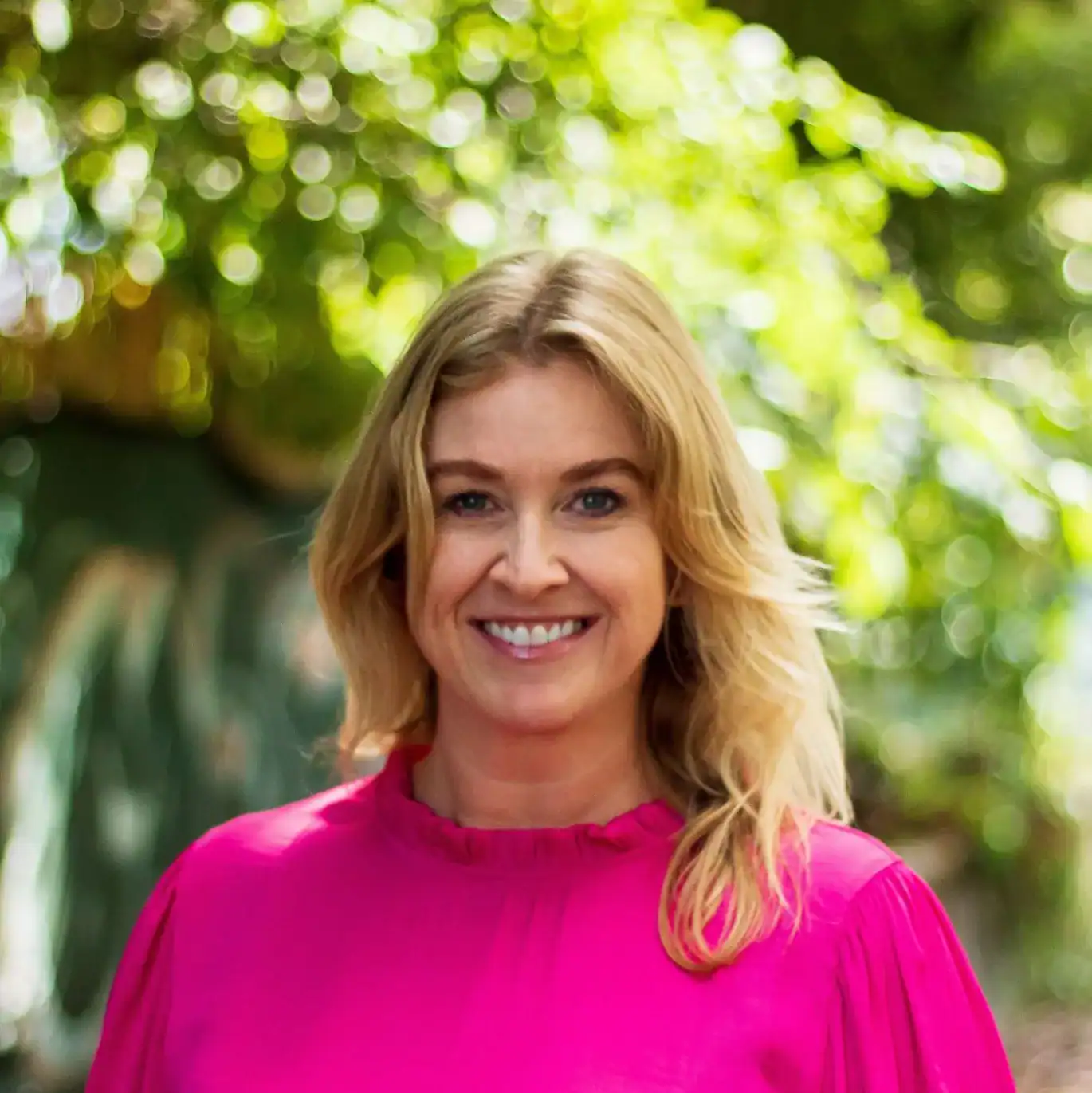 Vibrant woman smiling outdoors with sunlight filtering through green trees.