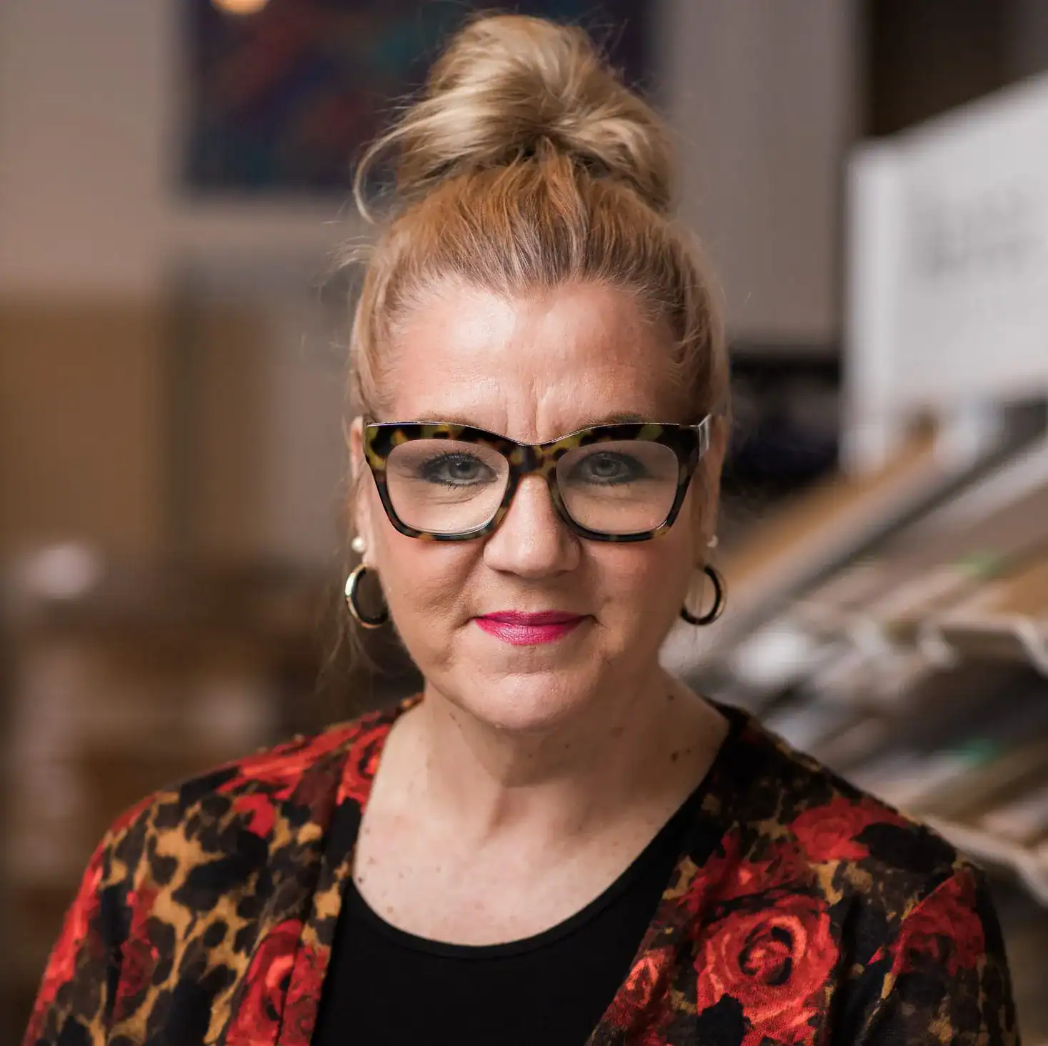 Close-up of a woman with red hair in a bun, wearing tortoiseshell glasses and a floral blazer, in a flooring showroom.