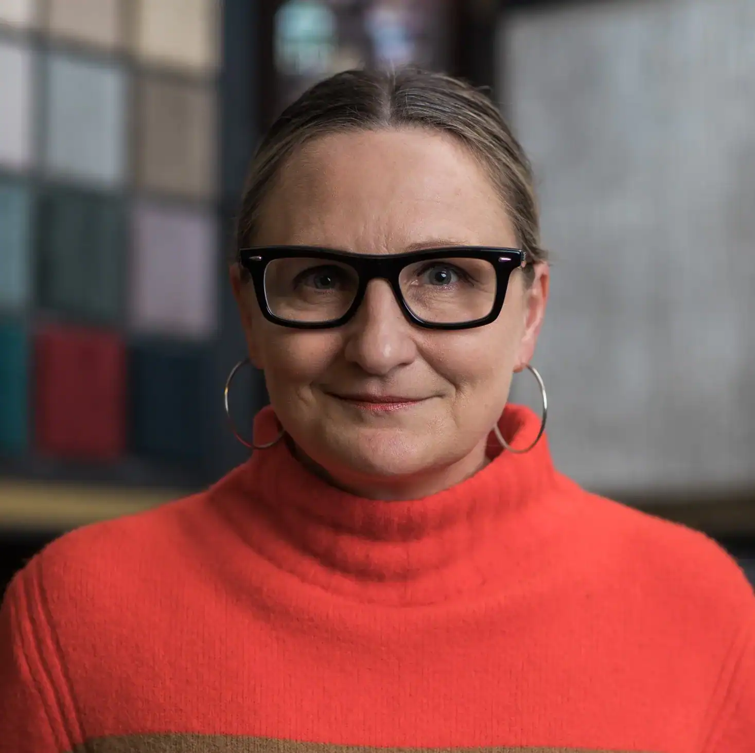 Close-up portrait of woman with glasses and earrings, stylish casual outfit in vibrant red turtleneck, modern urban background, professional indoor setting, confident expression, Dallas Watson Flooring.