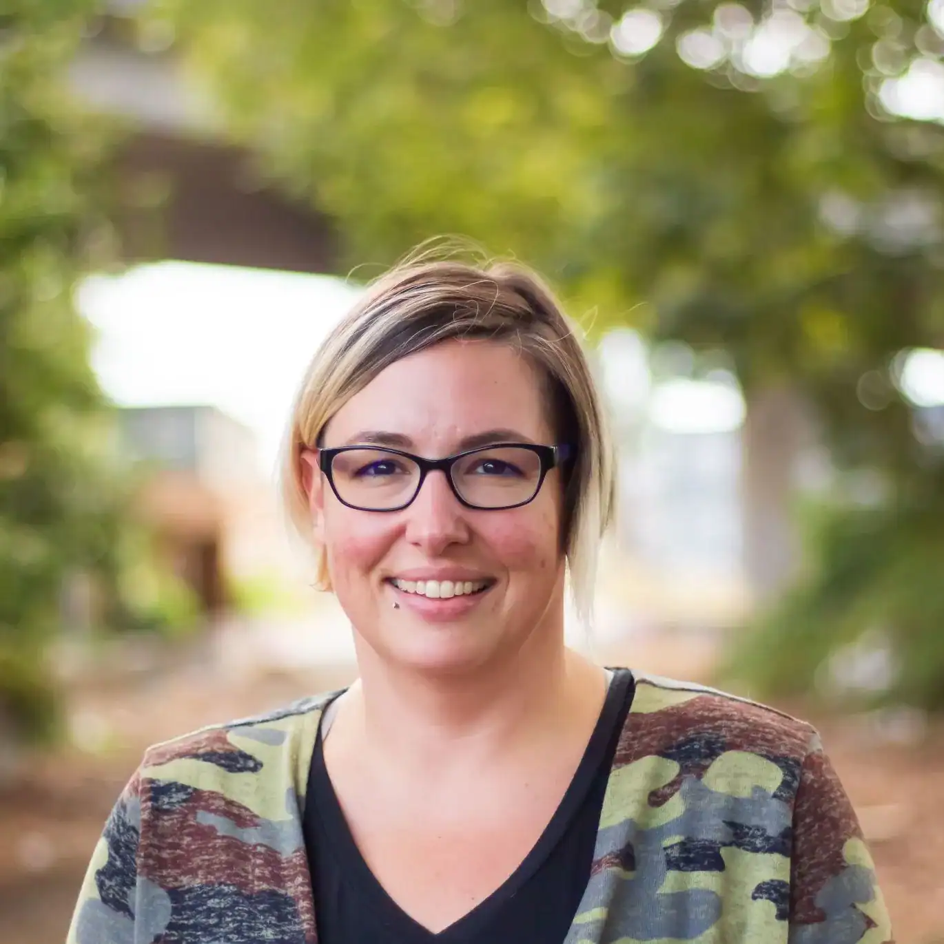 Woman with glasses smiling outdoors, trees background, casual attire, natural light, outdoor portrait, modern lifestyle, friendly expression.