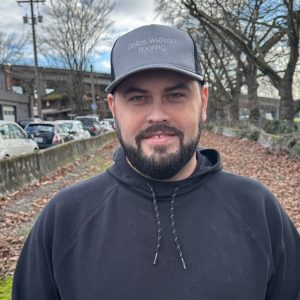 Men outdoors wearing black hoodie and gray cap with "dallas watson flooring" logo, standing on leaf-covered path in park, sunny day with trees and parked cars in background.