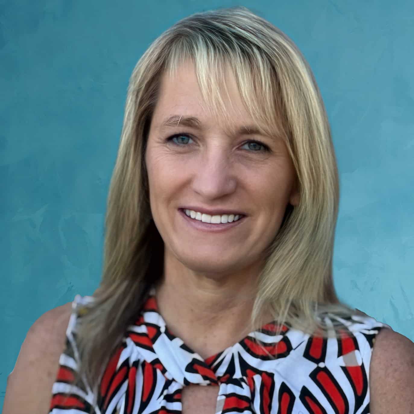 Close-up portrait of a smiling woman with blonde hair and blue eyes, wearing a patterned sleeveless top, against a blue background.